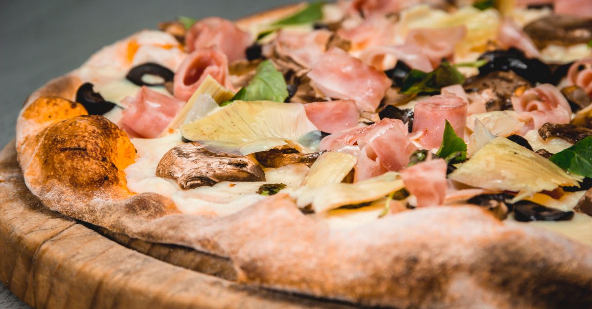 Nahaufnahme einer frisch gebackenen Pizza mit knusprigem Rand, Schinken, Oliven und Artischocken auf einem Holzbrett.