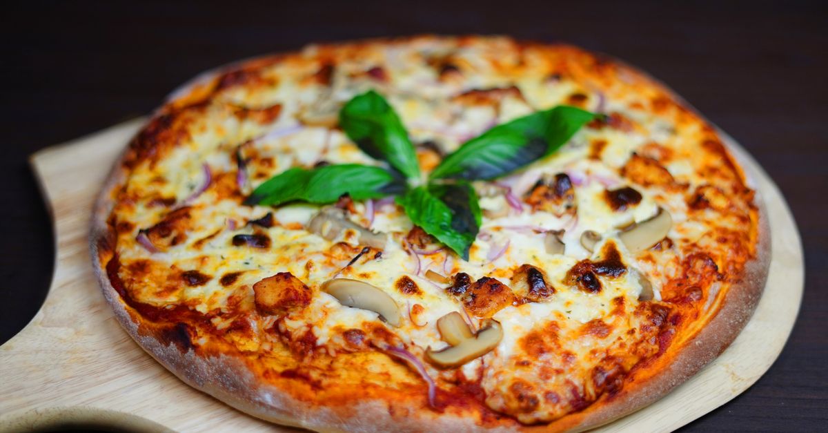 Eine ganze, knusprig gebackene Pizza mit Käse, Champignons und roten Zwiebeln liegt auf einem runden Holzbrett, garniert mit frischem Basilikum.