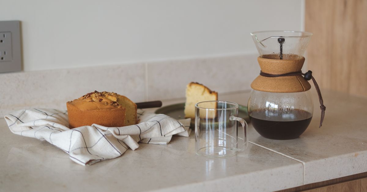 Ein Stück Kuchen und ein kleiner Kastenkuchen liegen auf einem Küchentuch neben einer leeren Glastasse und einer Kaffeekaraffe auf einer Küchenarbeitsplatte.