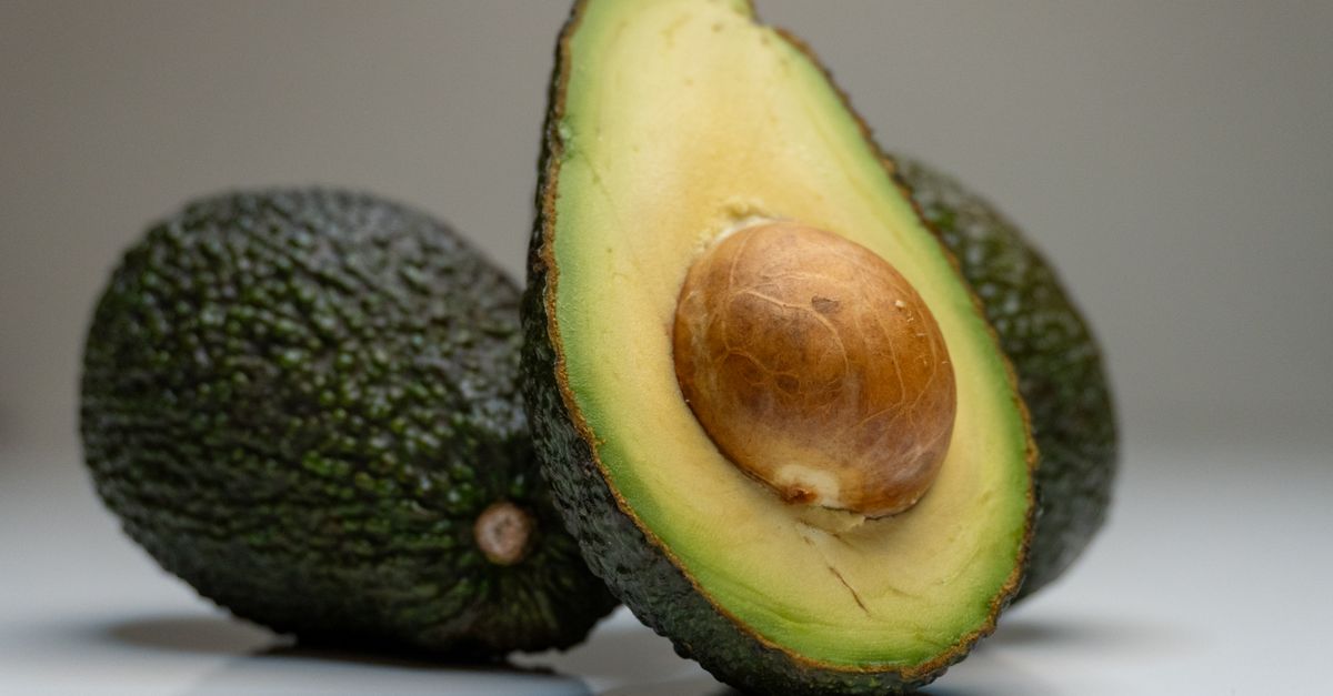 Eine halbierte Avocado mit sichtbarem Kern liegt vor einer weiteren ganzen Avocado.