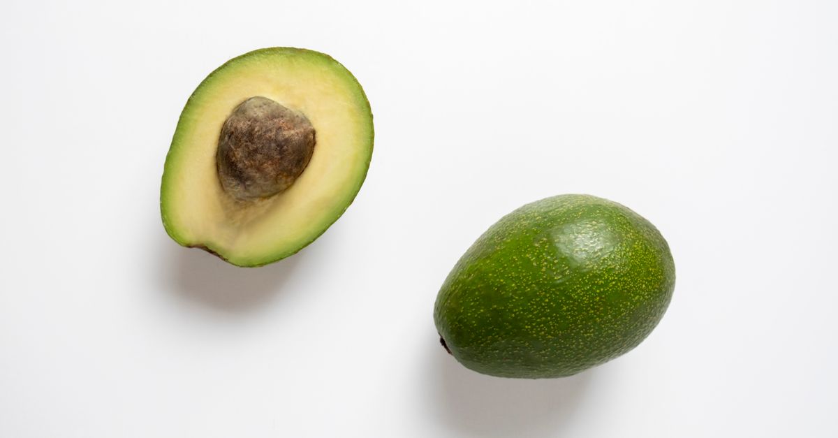 Eine halbierte Avocado mit Kern liegt neben einer ganzen Avocado auf weißem Untergrund.
