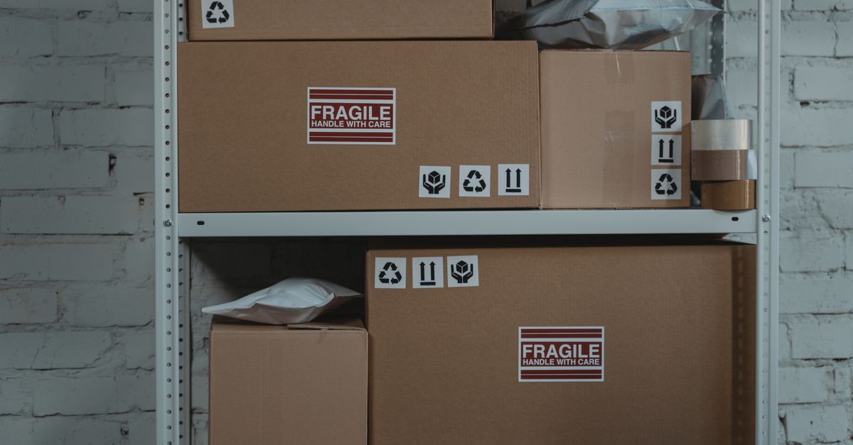 Ein Metallregal mit gestapelten Kartons, die mit „Fragile – Handle with care“ Etiketten und Symbolaufklebern versehen sind, vor einer weiß gestrichenen Backsteinwand.