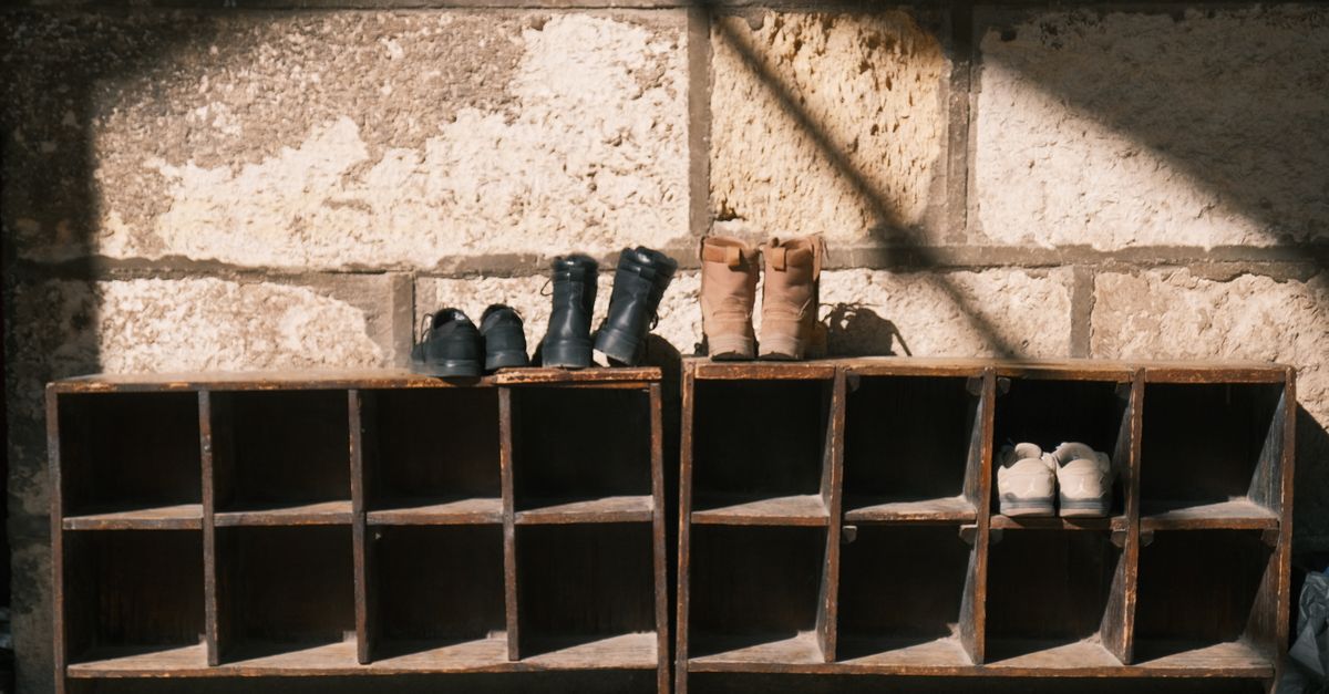 Zwei alte Holzregale mit offenen Fächern an einer Steinwand, auf denen einige Paar Schuhe und Stiefel stehen.