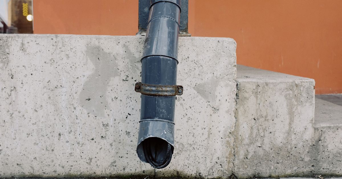 Ein graues Regenfallrohr führt an einer Betonmauer entlang nach unten neben einer Steintreppe.
