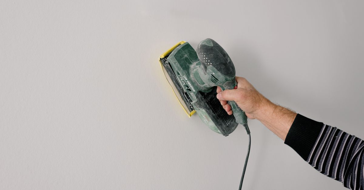 Eine Hand führt einen elektrischen Schwingschleifer über eine helle Wandfläche zum Glätten der Oberfläche.