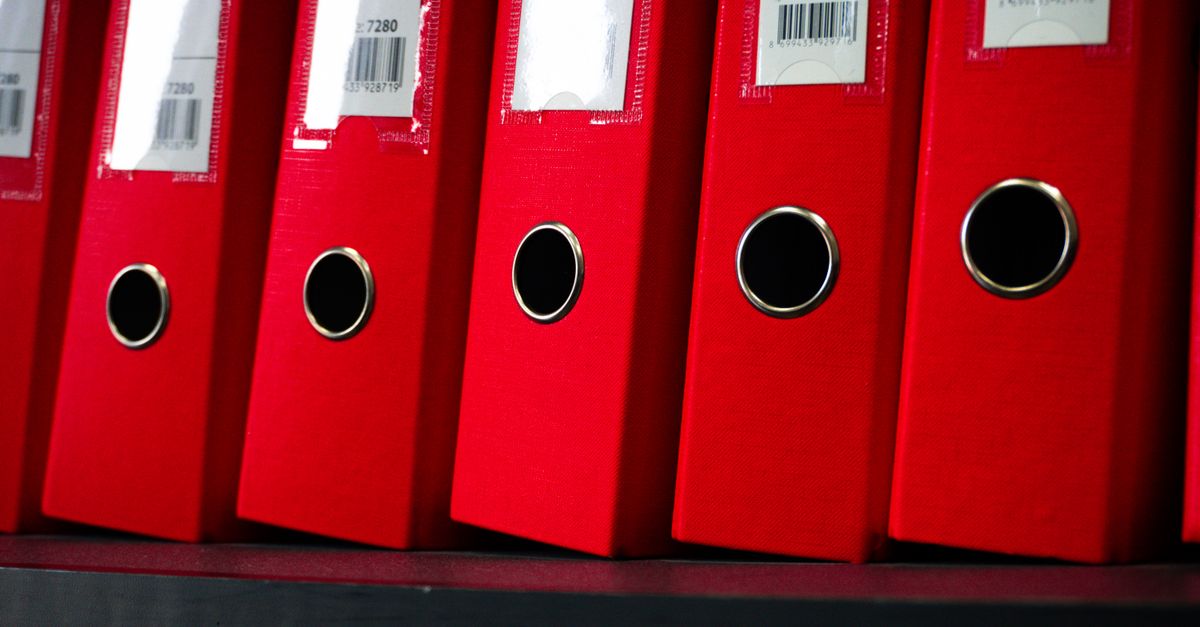 Reihe roter Aktenordner mit Beschriftungsschildern steht nebeneinander im Regal.