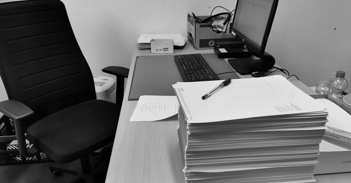 Schwarz-weiß Foto eines Schreibtisches mit einem ordentlich gestapelten Papierstapel, Stift, Computer und Bürostuhl im Hintergrund.
