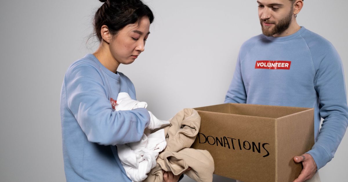 Eine Frau sortiert Kleidung in einen Karton mit der Aufschrift 'Donations', während ein Mann mit Volunteer-Pullover den Karton hält.