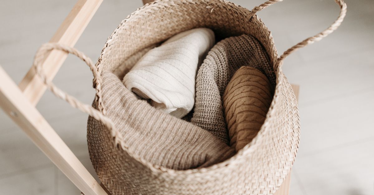 Ein geflochtener Korb mit ordentlich gerollten Strickpullovern in Beige- und Naturtönen steht auf einem Holzgestell.