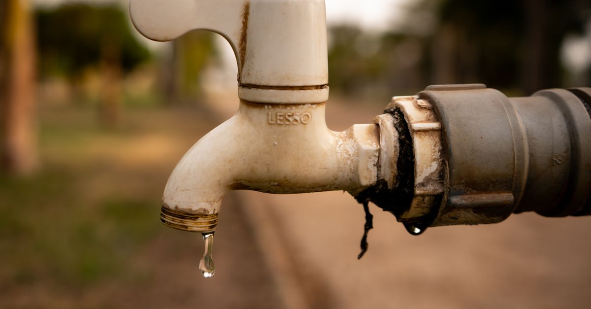 Nahaufnahme eines tropfenden Wasserhahns mit Kalkablagerungen und rostigen Verschraubungen im Freien.