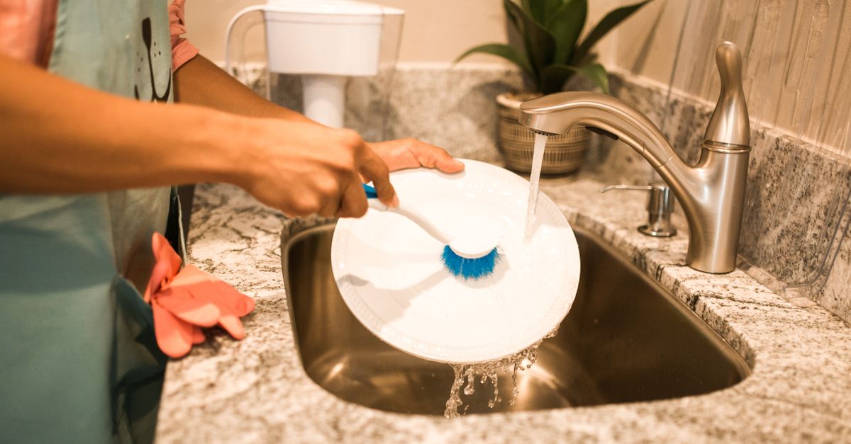 Eine Person spült mit einer blauen Bürste einen Teller unter fließendem Wasser an einer Küchenspüle mit Steinarbeitsplatte.