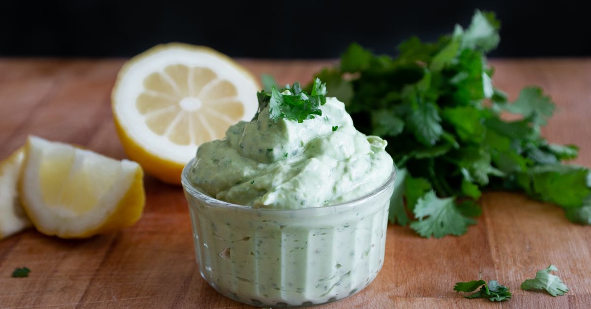 Eine cremige, hellgrüne Sauce mit frischen Kräutern in einer Glasschale, daneben liegen eine halbierte und eine geviertelte Zitrone auf einem Holzbrett.