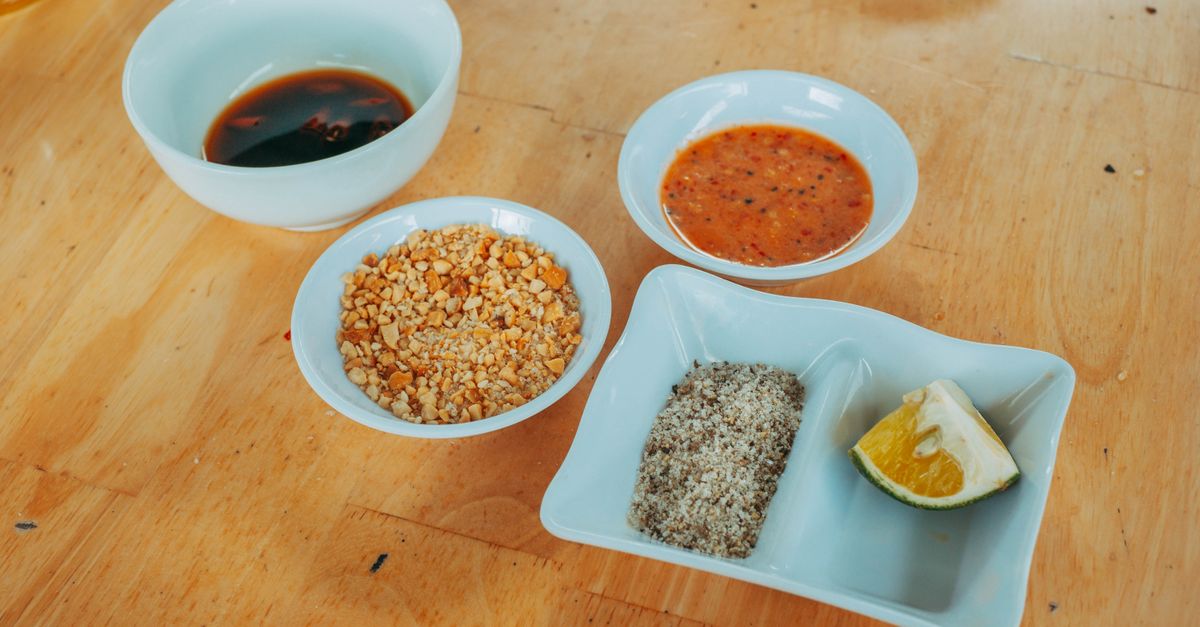 Mehrere kleine weiße Schälchen mit unterschiedlichen Saucen, darunter dunkle Sojasauce, rote Chilisauce, gehackte Erdnüsse und eine Gewürzmischung mit Limettenspalte auf einem Holztisch.
