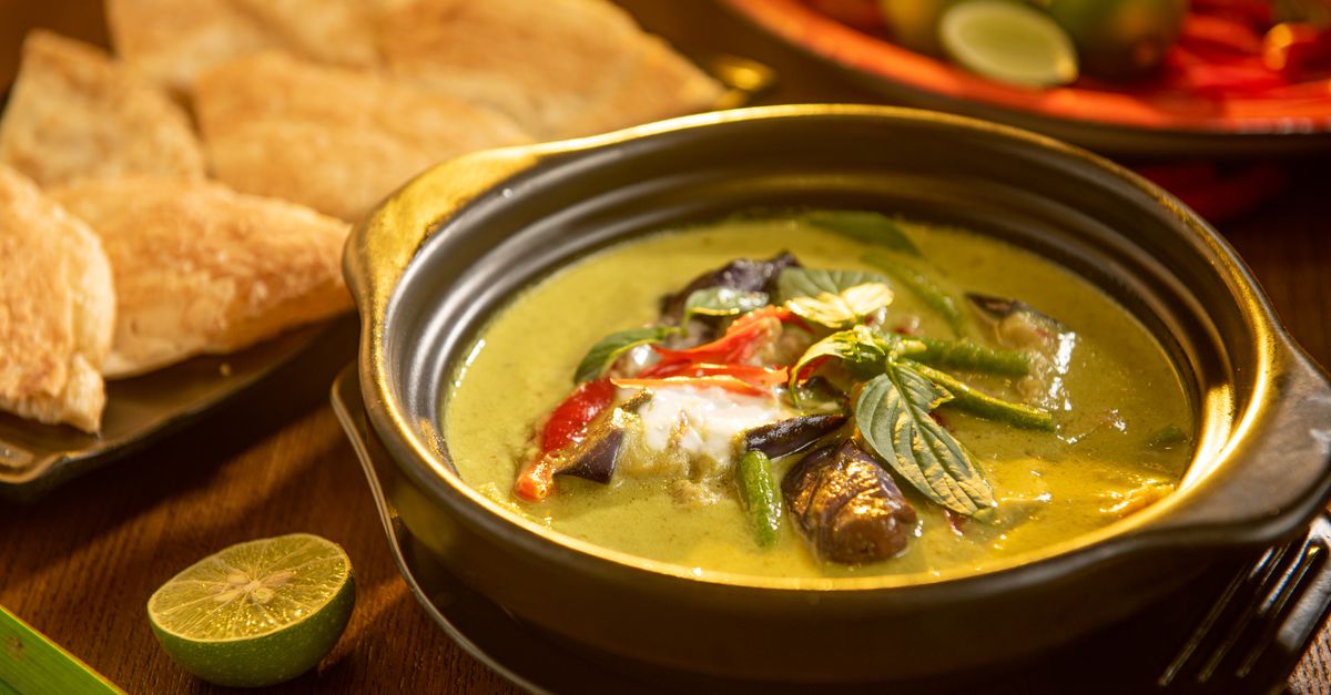 Eine grüne Curry-Suppe mit Kokosmilch, Auberginen, Bohnen und Kräutern in einer schwarzen Schale, daneben Limette und Fladenbrot.