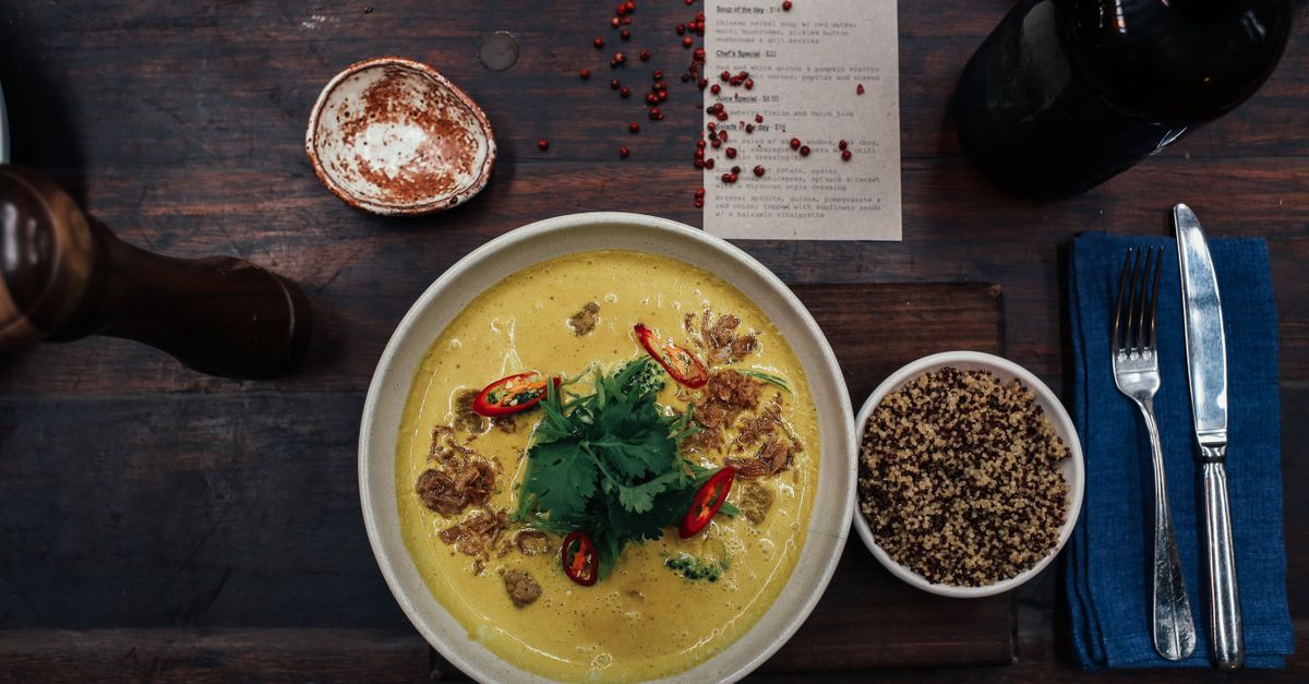 Ein gelbes Curry mit roten Chilistücken und Koriander in einer Schüssel auf einem dunklen Holztisch, daneben eine Schale mit Quinoa und Besteck.
