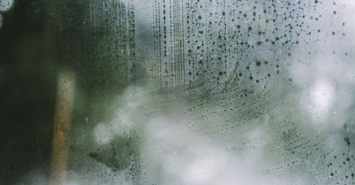 Beschlagene Fensterscheibe mit herablaufenden Kondenswassertropfen, im Hintergrund verschwommen Bäume und Licht zu erkennen.