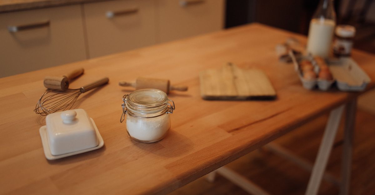 Eine Butterdose mit Butter, ein Schneebesen, ein Glas mit Zucker, ein Nudelholz und ein Eierkarton stehen auf einem Holztisch in der Küche.