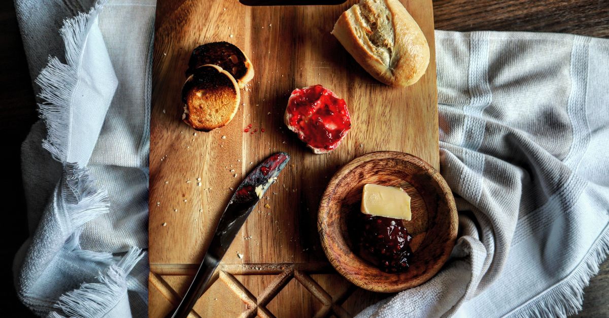 Auf einem Holzbrett liegt ein kleines Schälchen mit einem Stück weicher, gestrichener Butter neben Marmelade und angeschnittenem Brot.