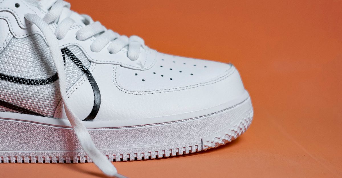 Nahaufnahme eines weißen Sneakers mit schwarzem Logo-Streifen vor orangefarbenem Hintergrund.