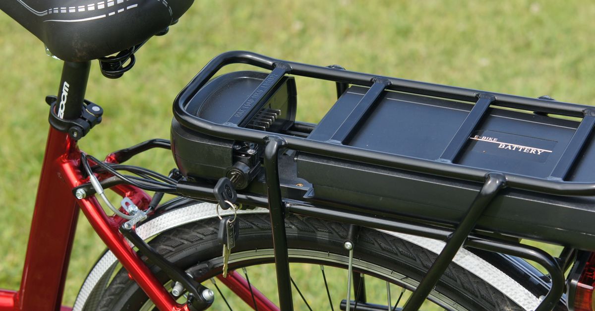 Nahaufnahme eines schwarzen E-Bike-Akkus auf dem Gepäckträger eines roten Fahrrads im Gras.