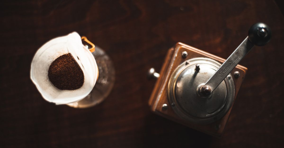Gemahlener Kaffee in einem Papierfilter neben einer alten Handkaffeemühle auf einem dunklen Holztisch, von oben fotografiert.