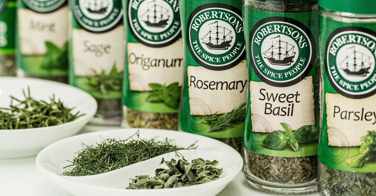 Mehrere Gewürzgläser mit Etiketten wie Rosemary, Sweet Basil und Sage stehen in einer Reihe neben weißen Schälchen mit getrockneten Kräutern.