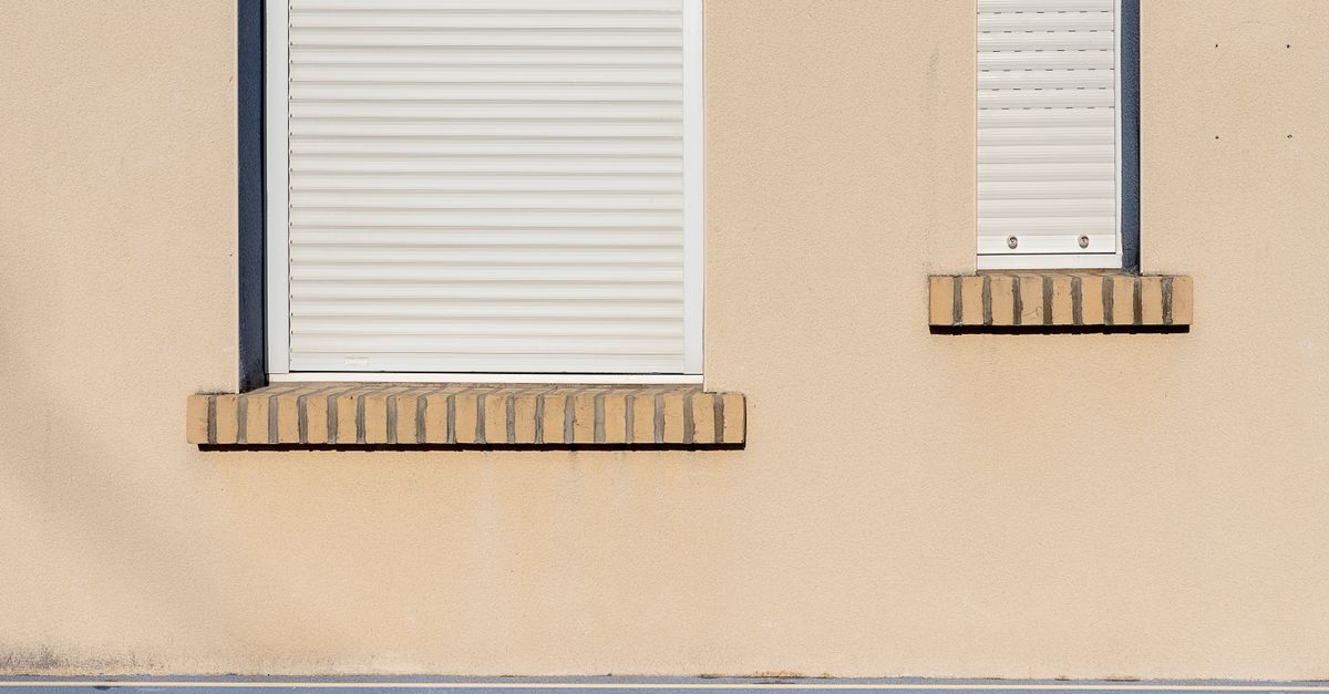 Fassade eines Wohnhauses mit zwei heruntergelassenen weißen Rollläden über den Fenstern am Nachmittag.