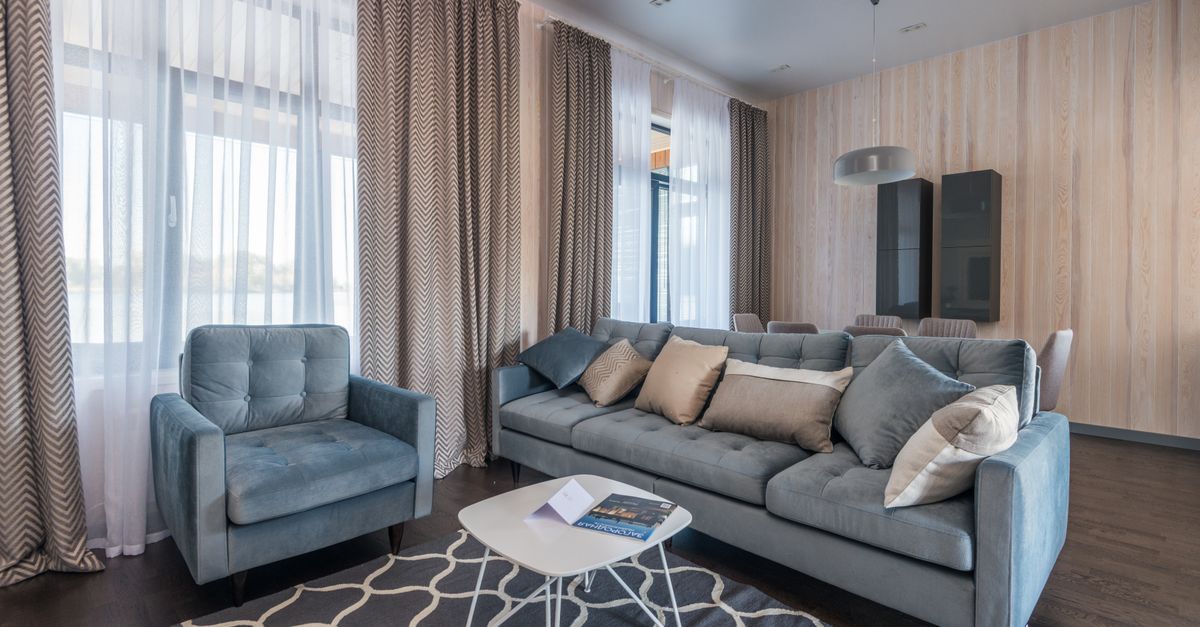 Wohnzimmer mit dicken Vorhängen an großen Fenstern, einem Teppich und gepolsterten Sofas, die zur Schalldämmung beitragen können.