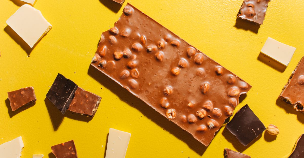 Zerbrochene Schokoladentafel mit Haselnüssen auf gelbem Hintergrund, umgeben von Stücken weißer und dunkler Schokolade.