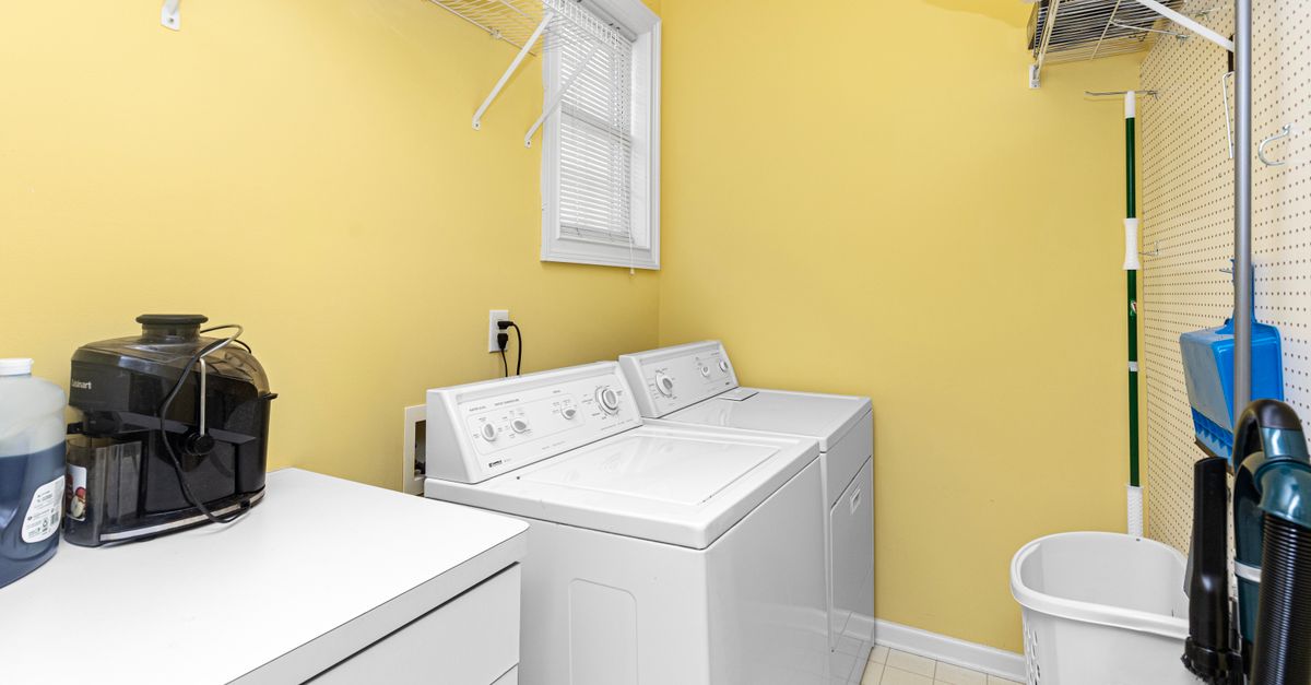 Ein heller Hauswirtschaftsraum mit gelben Wänden, Waschmaschine und Trockner nebeneinander sowie einem Ablagesystem und Regalen an der Decke.