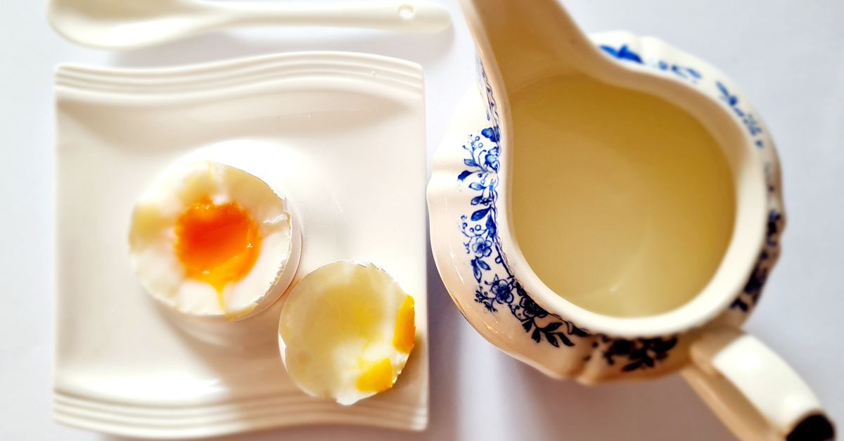 Ein weichgekochtes Ei im Eierbecher, aufgeschnitten mit flüssigem Eigelb, daneben eine Kanne und ein Löffel auf dem Frühstückstisch.