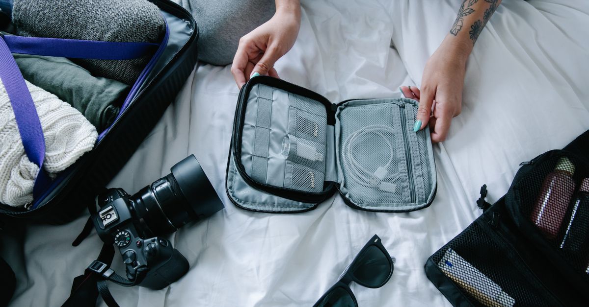Draufsicht auf ein aufgeschlagenes Kabel-Organizer-Etui mit Ladekabeln neben Kamera, Sonnenbrille und einem Koffer voller gefalteter Kleidung auf einem Bett.