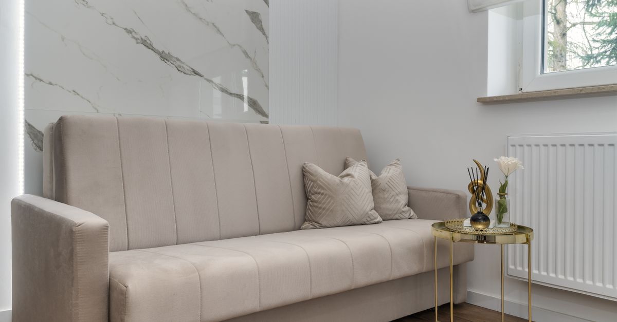 Ein weißer Heizkörper unter einem Fenster neben einem beigefarbenen Sofa in einem Wohnzimmer mit Holzfußboden.