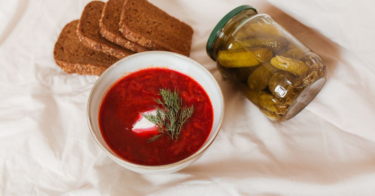 Eine Schüssel roter Borschtsch mit Sauerrahm und Dill, daneben Roggenbrotscheiben und ein Glas eingelegte Gurken.