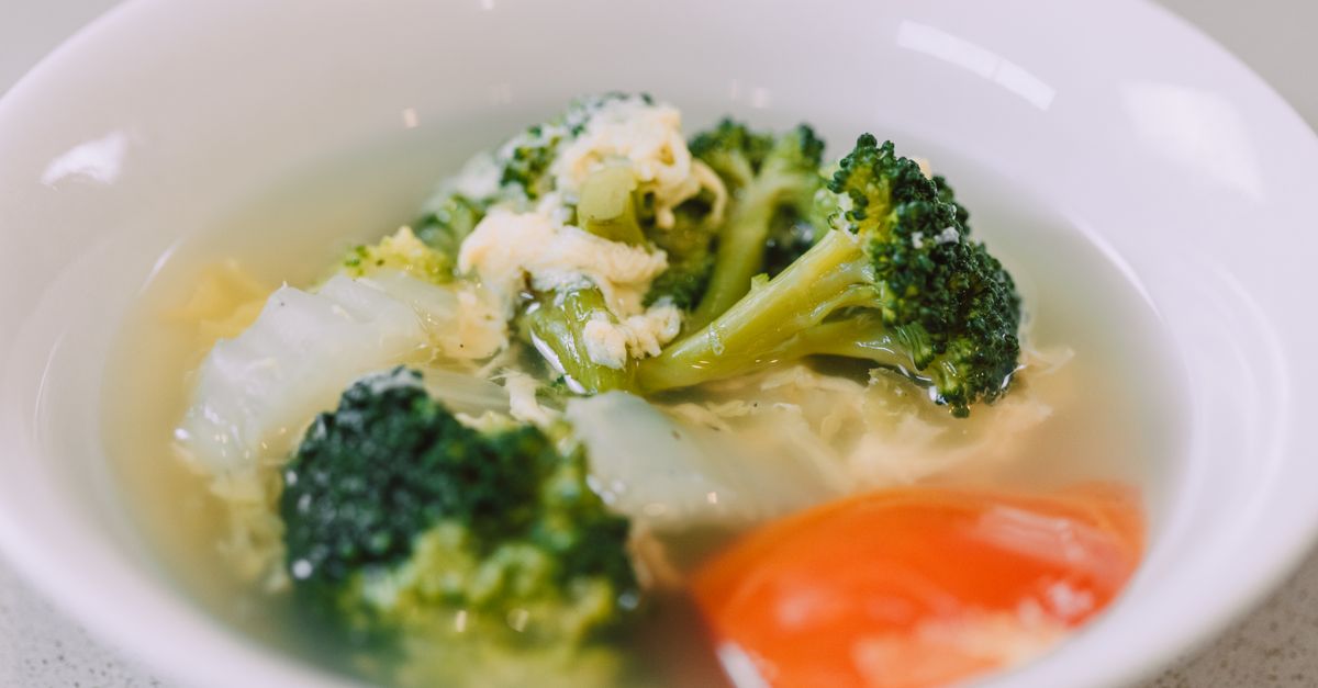 Eine weiße Schale mit klarer Gemüsesuppe, gefüllt mit Brokkoli, Kohl und einem Stück Tomate.