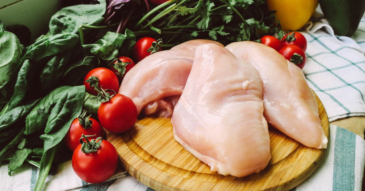 Rohe Hähnchenbrustfilets liegen auf einem runden Holzbrett, umgeben von Kirschtomaten und frischem Blattgemüse.