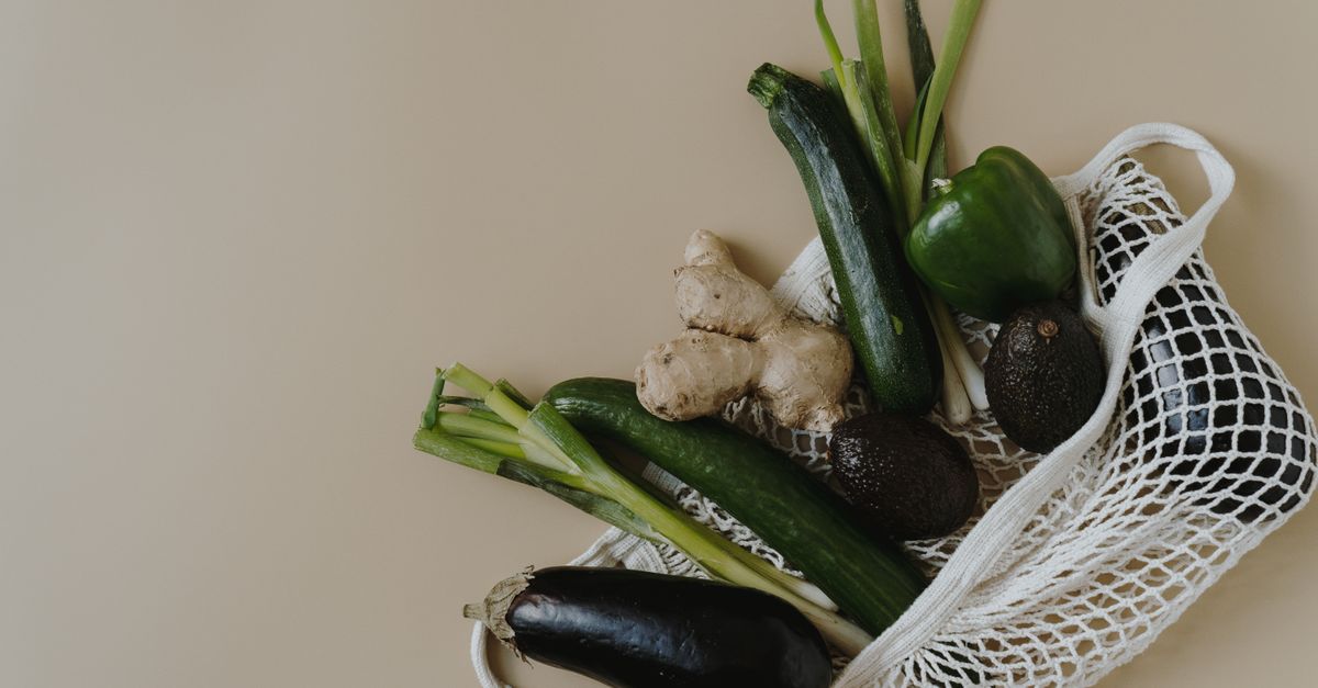 Ein Einkaufsnetz mit Zucchini, Gurken, Ingwer, Paprika, Frühlingszwiebeln und Auberginen liegt auf einer beigen Fläche.
