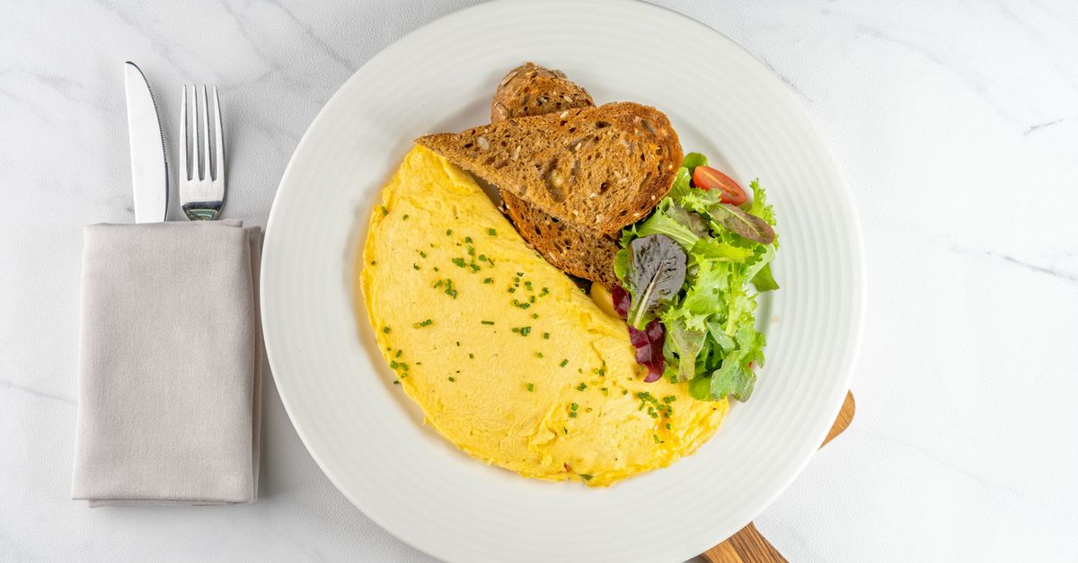 Ein gefaltetes Omelett mit Kräutern liegt auf einem weißen Teller neben einer Scheibe Vollkornbrot und einem kleinen Blattsalat.