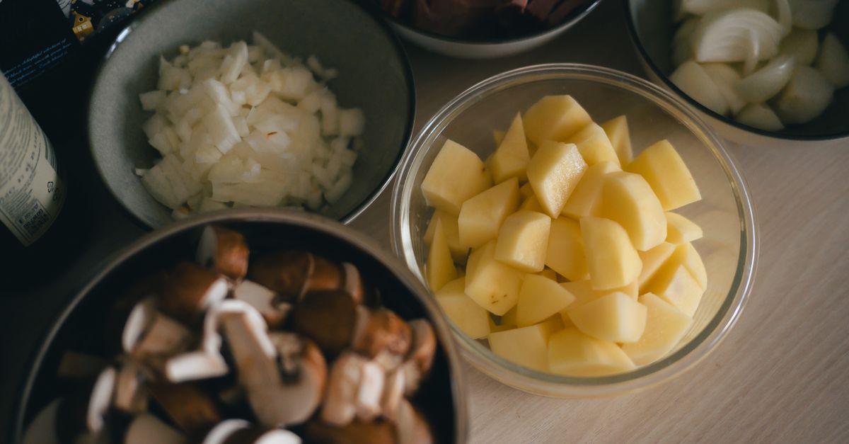 Eine Glasschüssel mit gewürfelten rohen Kartoffeln steht neben Schüsseln mit gehackten Zwiebeln und Pilzen auf einem Tisch.