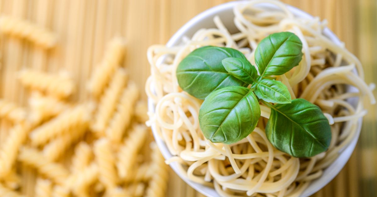 Eine weiße Schale mit gekochten Spaghetti und frischem Basilikum, daneben liegen ungekochte Fusilli auf einem Holzbrett.