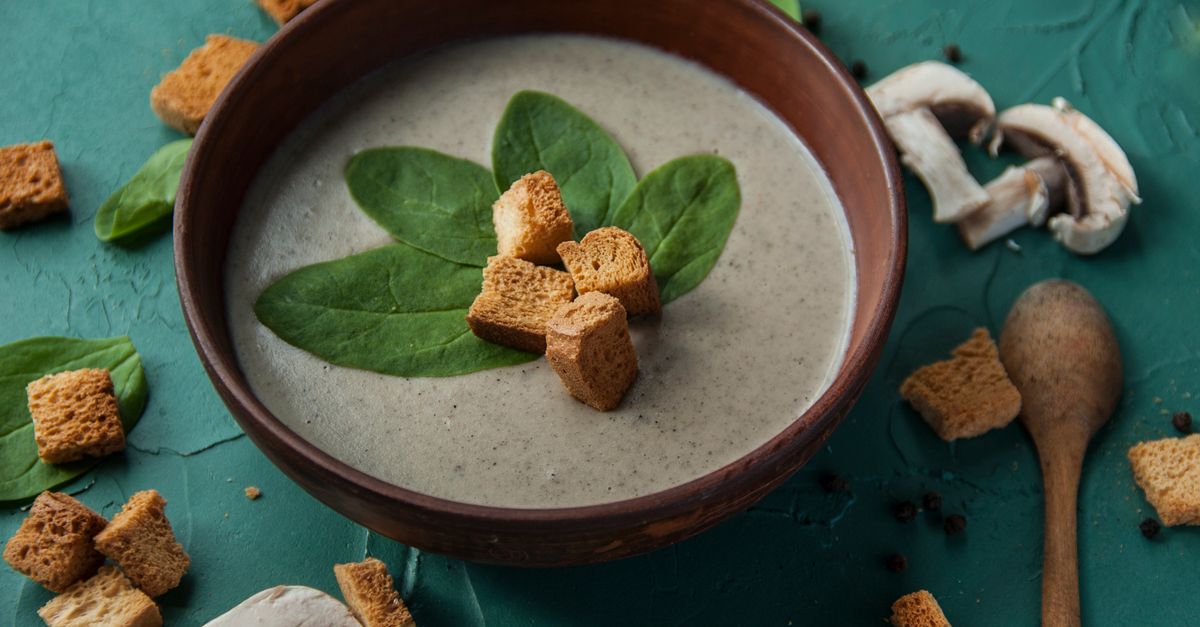 Eine cremige Pilzsuppe in einer rustikalen Keramikschale, garniert mit Spinatblättern und knusprigen Croutons, umgeben von frischen Champignonscheiben.