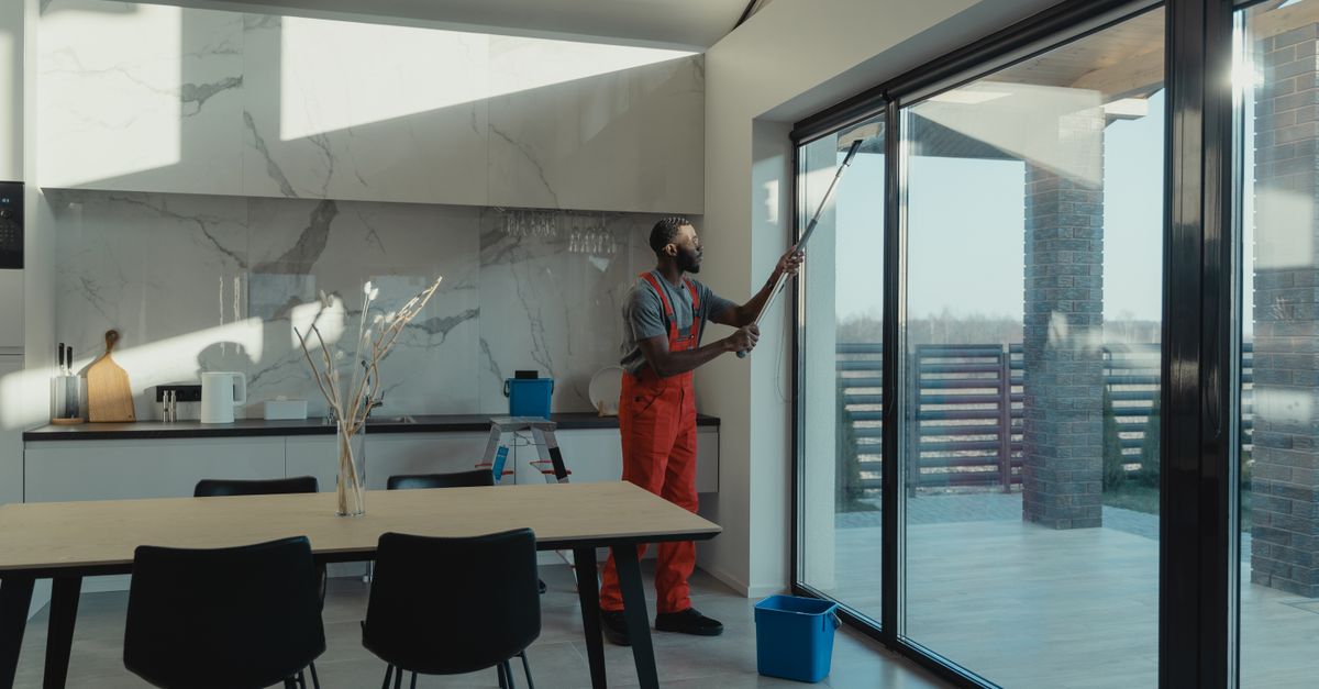 Ein Mann in Arbeitskleidung reinigt mit einem Fensterwischer eine große Glasschiebetür in einem modernen Wohnraum.