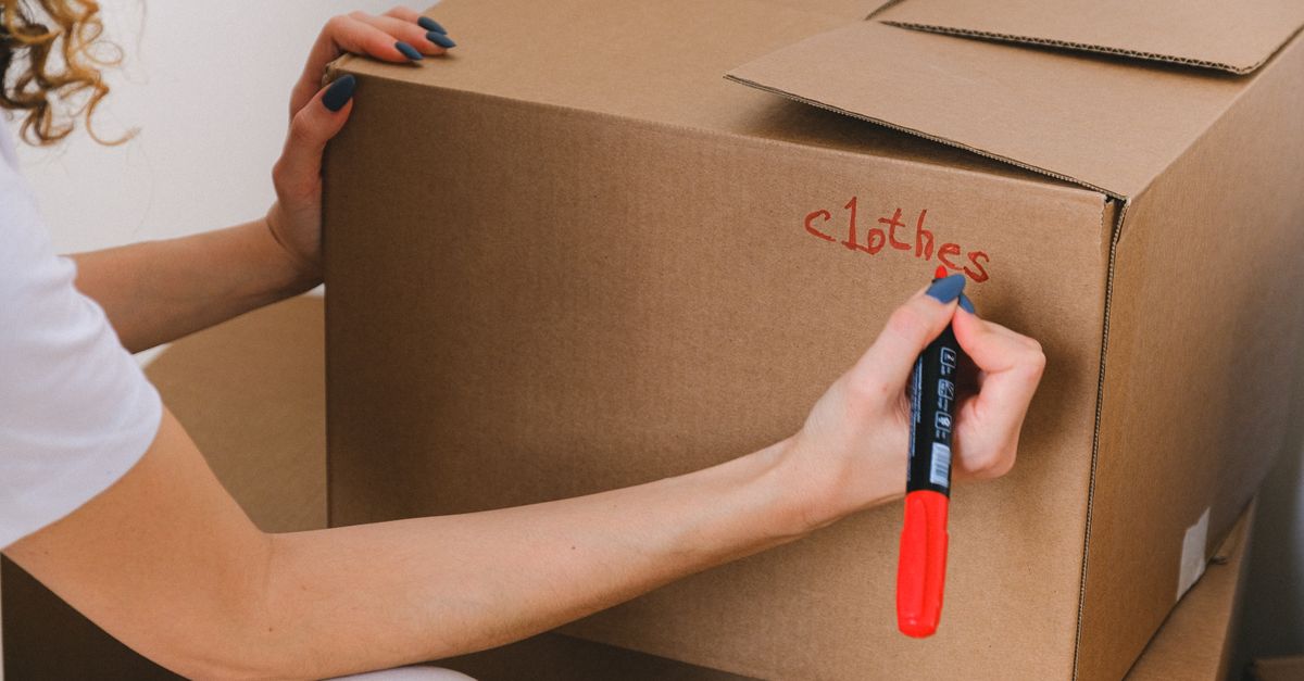 Eine Frau beschriftet einen Umzugskarton mit rotem Marker mit dem Wort 'clothes'.