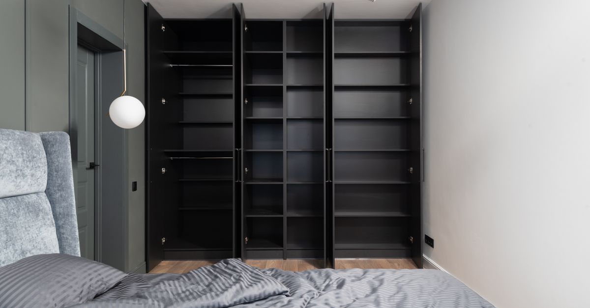 Ein offener, leerer Einbauschrank mit mehreren Fächern und Kleiderstange neben einem Bett im Schlafzimmer.