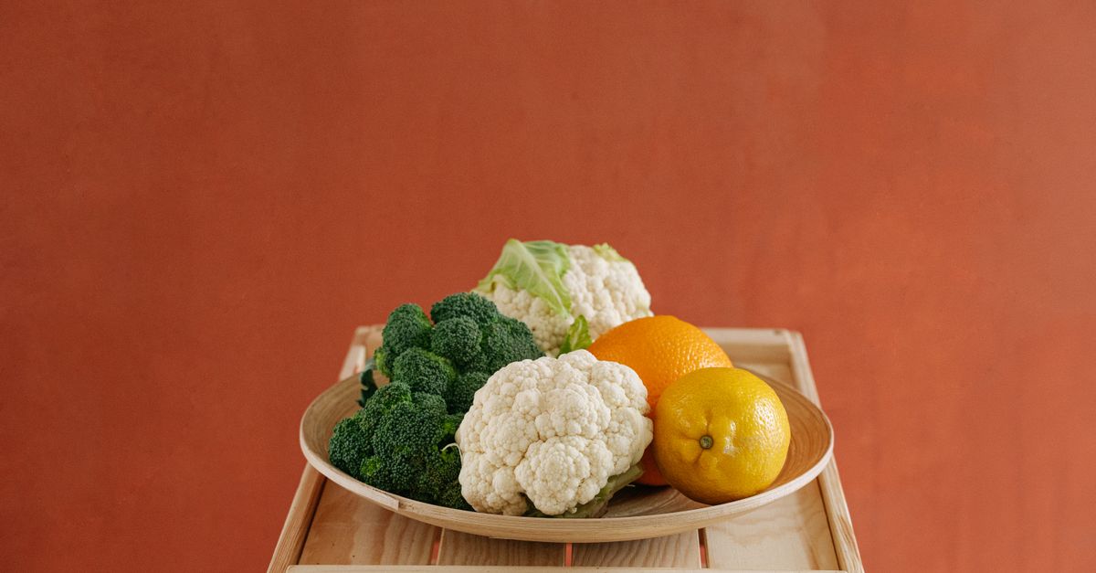 Frischer Brokkoli und Blumenkohl liegen zusammen mit einer Orange und einer Zitrone auf einem Holzteller vor orangefarbenem Hintergrund.