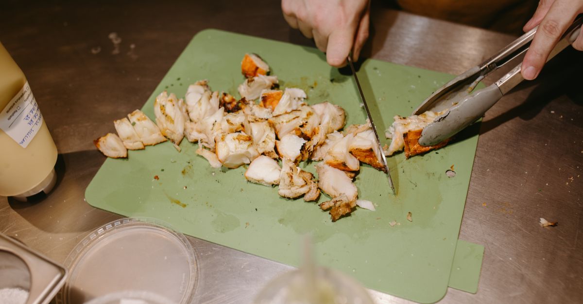 Auf einem grünen Schneidebrett wird gekochtes, in Stücke geschnittenes Hühnerfleisch mit Messer und Zange zerkleinert.