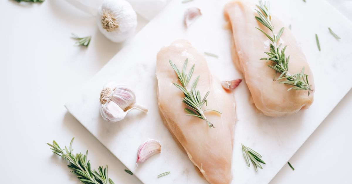 Zwei rohe Hühnerbrustfilets mit Rosmarinzweigen auf einem weißen Marmorbrett, umgeben von Knoblauchknollen.