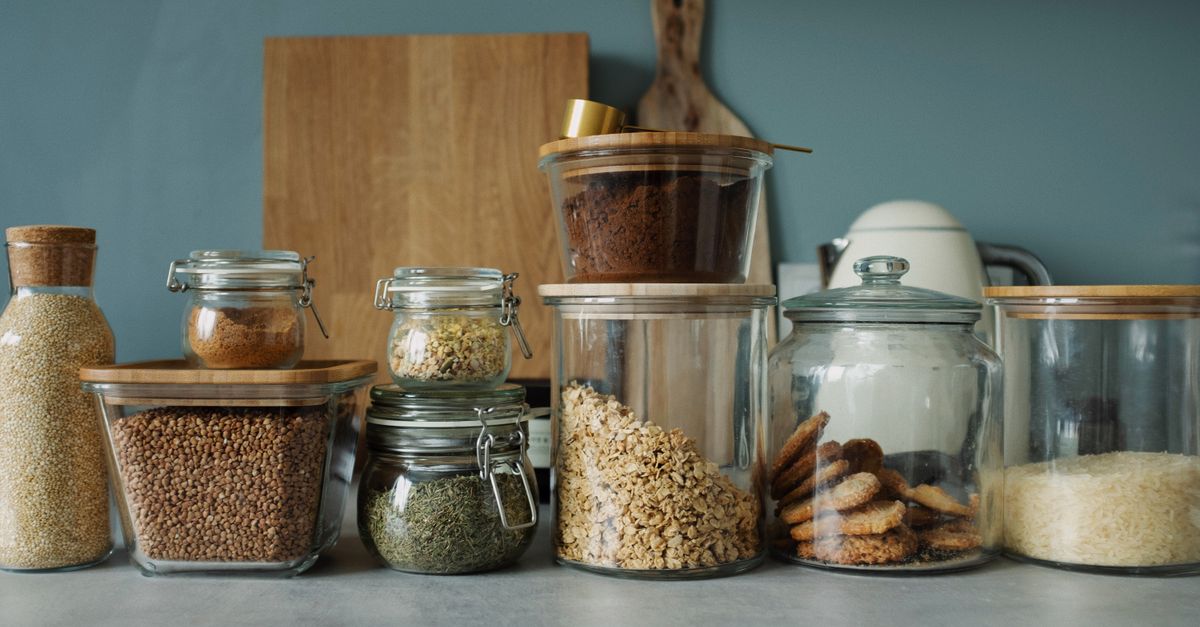 Verschiedene Vorratsgläser mit Müsli, Getreide, Keksen und Reis stehen aufgereiht auf einer Küchentheke.