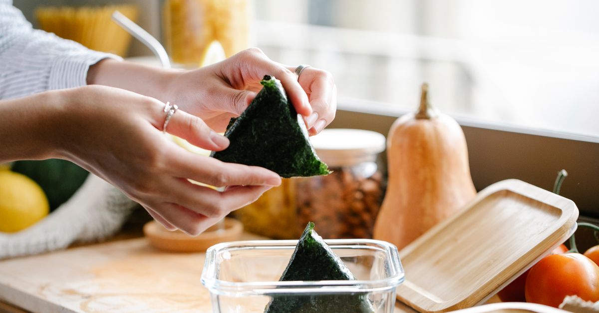 Zwei Hände falten ein dreieckiges Nori-Onigiri über einer Frischhaltebox, im Hintergrund liegen Gemüse und ein Holzbrett auf der Küchenarbeitsplatte.