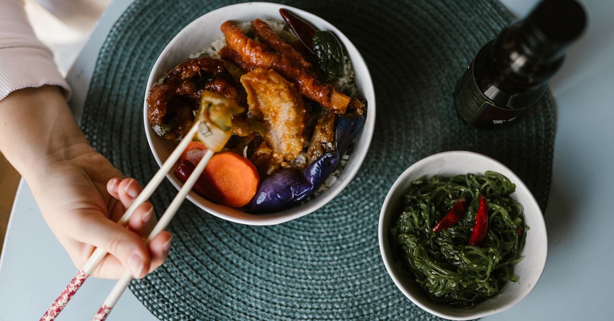 Draufsicht auf einen Tisch mit einer Schale mariniertem grünem Wakame-Algensalat neben einer Schale mit Reis, Fleisch und Gemüse, während eine Hand mit Stäbchen danach greift.